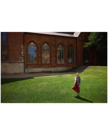 Young woman stands on the green lawn outside the red brick building, wearing a plaid shirt and long red skirt, on a sunny day.
