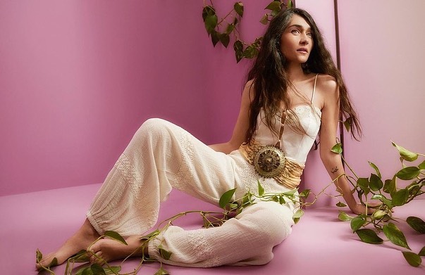 Portrait of a woman with long hair wearing a white outfit with a statement pendant, surrounded by green leafy vines against a pink backdrop.