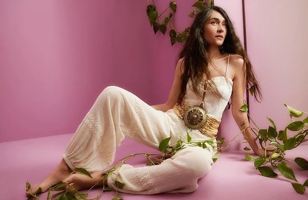 Portrait of a woman with long hair wearing a white outfit with a statement pendant, surrounded by green leafy vines against a pink backdrop.