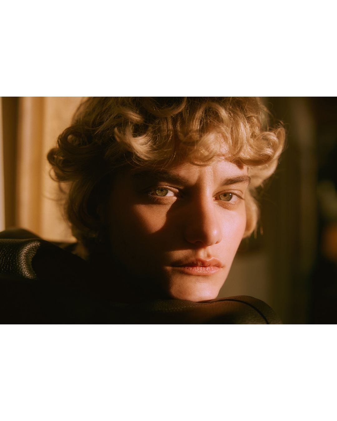 Close-up portrait of a blonde adult female with short curly hair looking directly at the camera, illuminated with golden-hour lighting indoors.