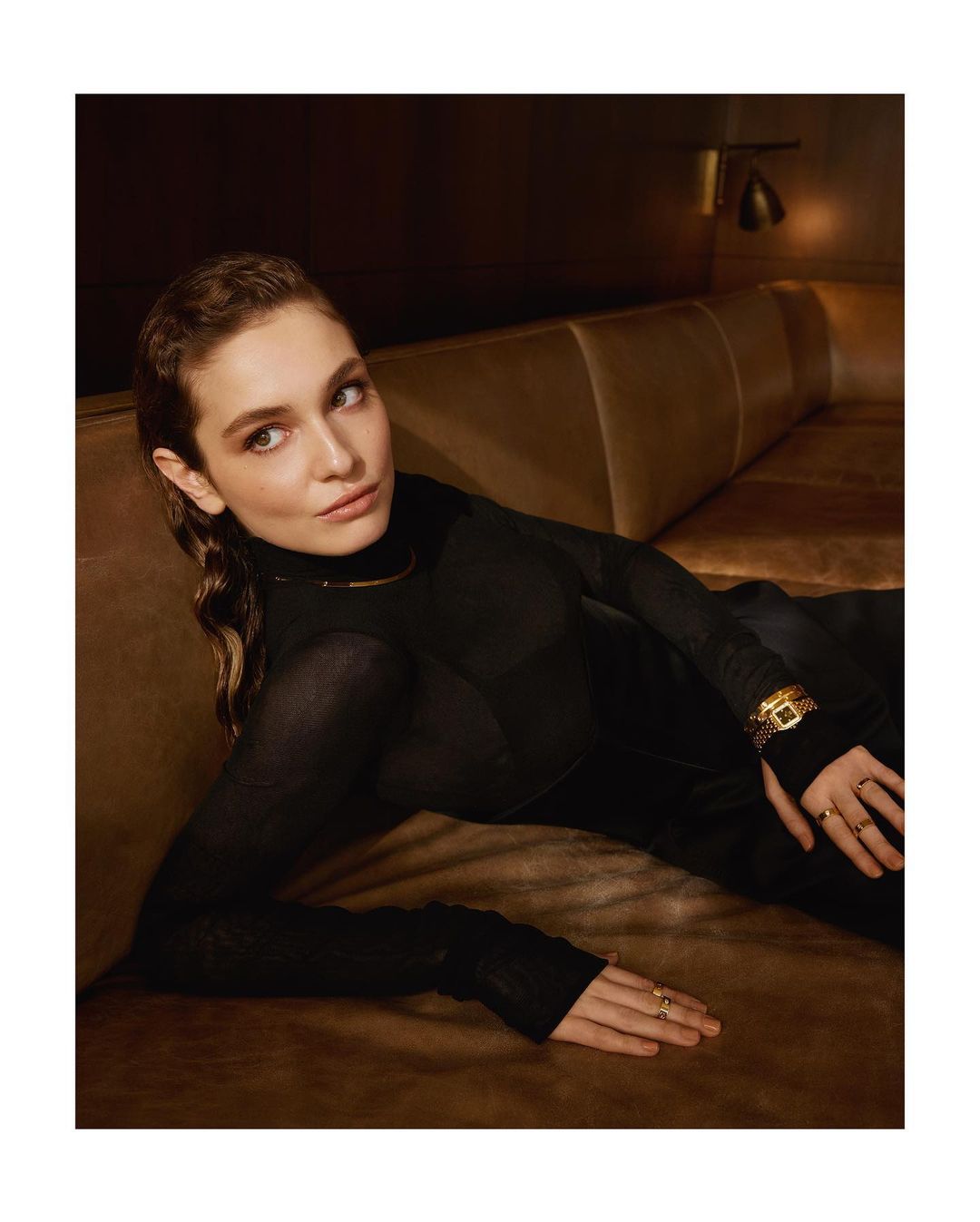 Adult woman with long, wavy brown hair poses on a brown leather sofa. She wears black clothing and gold jewelry in a warm, indoor setting.