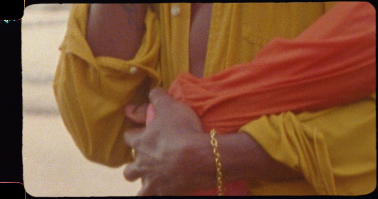 Close-up view of two people embracing, one in a mustard-yellow shirt and the other in an orange shirt, against a neutral backdrop.