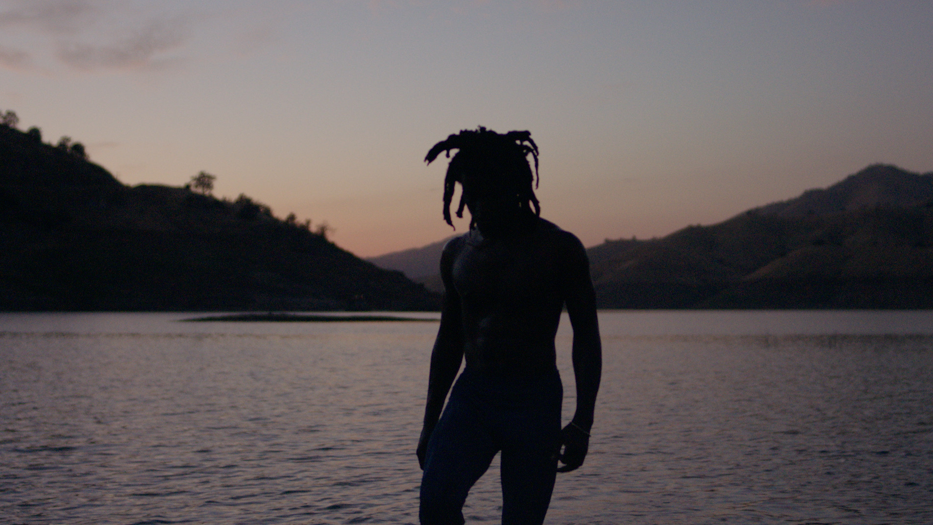 Silhouette of a shirtless adult male with dreadlocks standing in front of a lake at dusk, surrounded by distant mountains.