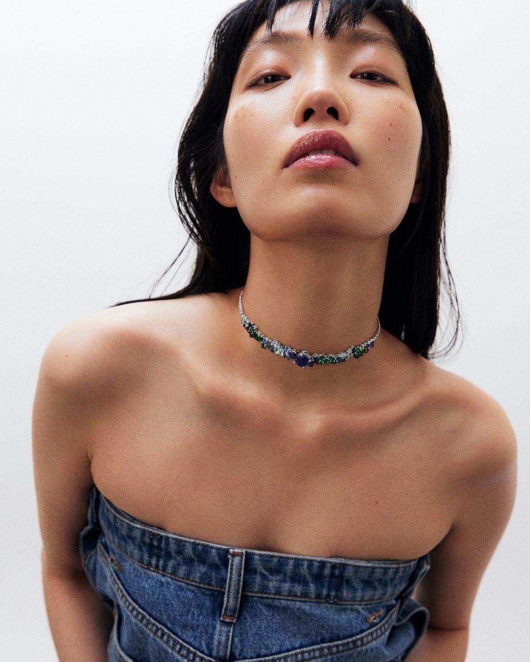 Woman with dark hair and denim top wearing a blue and green floral gemstone choker against a white background in a close-up portrait.