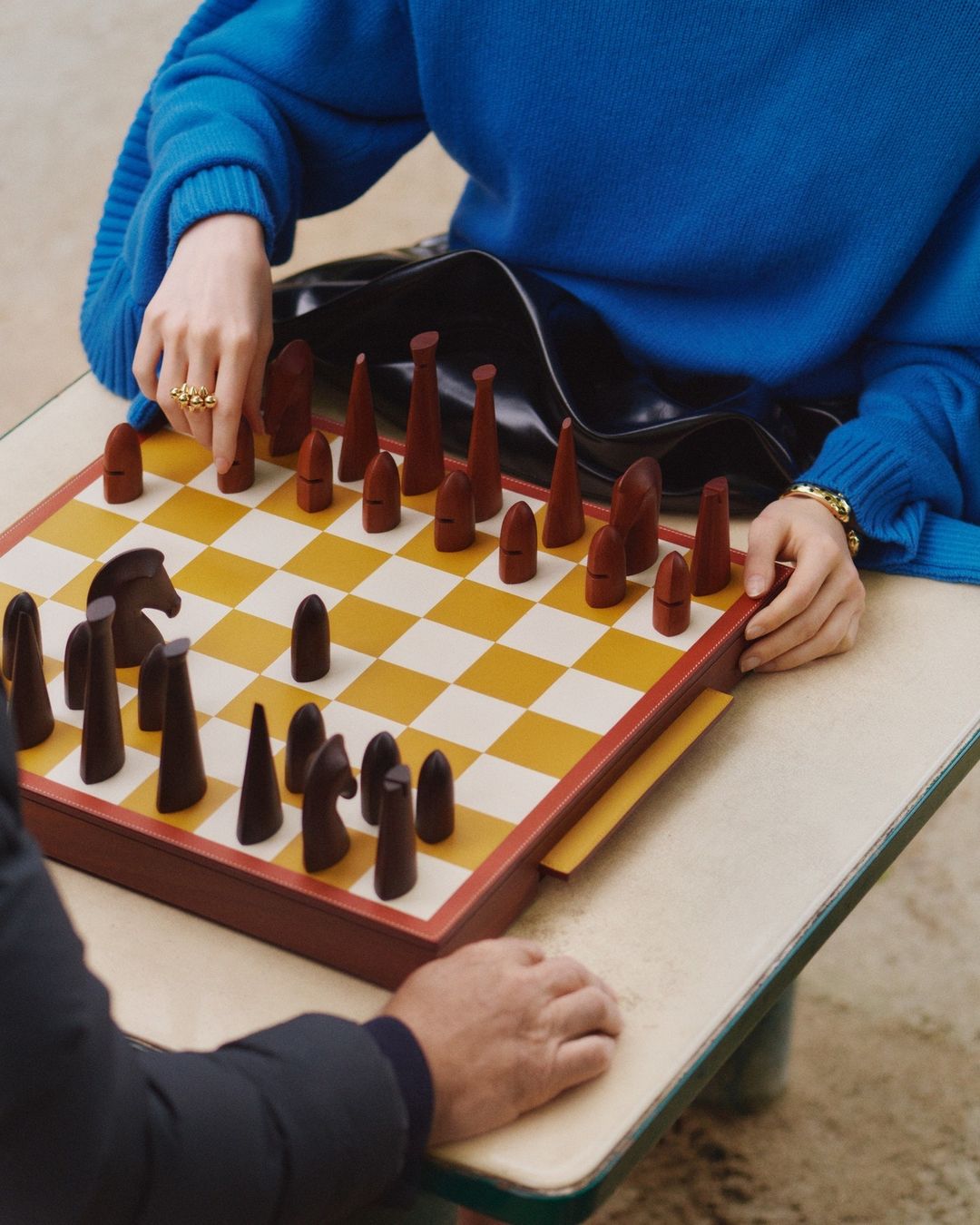 Two people playing chess on a checkered board. A woman wearing a blue sweater and a ring is visible along with her opponent.