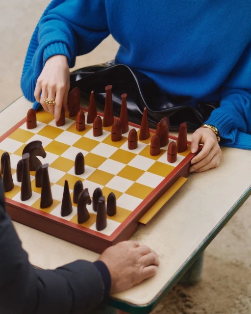 Two people playing chess on a checkered board. A woman wearing a blue sweater and a ring is visible along with her opponent.