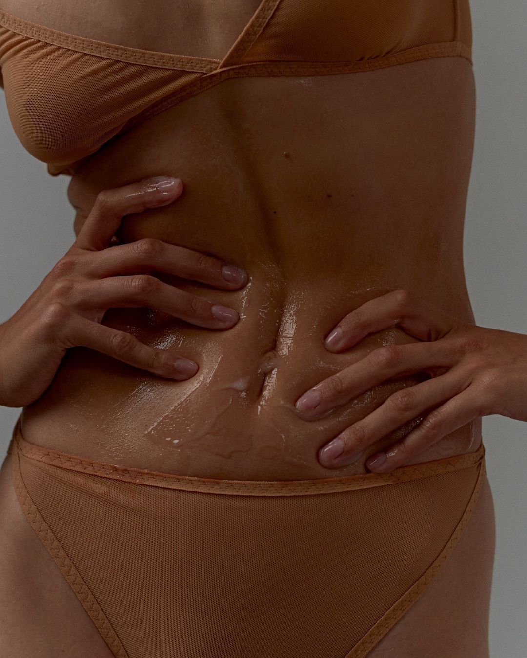 Close-up of a tanned adult female torso and hands with glossy sheen; subject is wearing a beige two-piece set against a grey studio backdrop.
