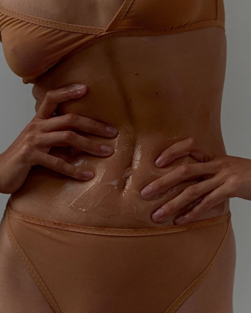 Close-up of a tanned adult female torso and hands with glossy sheen; subject is wearing a beige two-piece set against a grey studio backdrop.