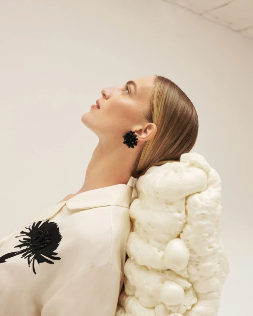 Model poses with a sculptural white form. She wears black flower earrings and a white collared shirt with a black flower design.