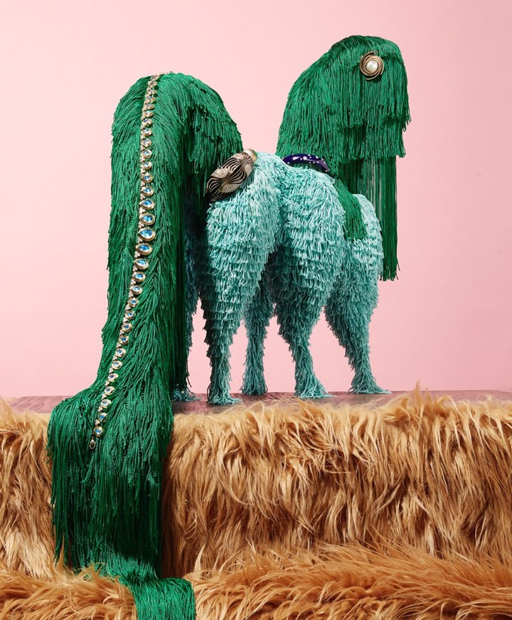 Two animal sculptures covered in fringes in shades of green and blue on a faux fur surface against a light pink background.