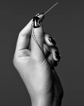 Close-up shot of a hand holding a needle with thread, showcasing spike-adorned black nail polish and a gray backdrop.