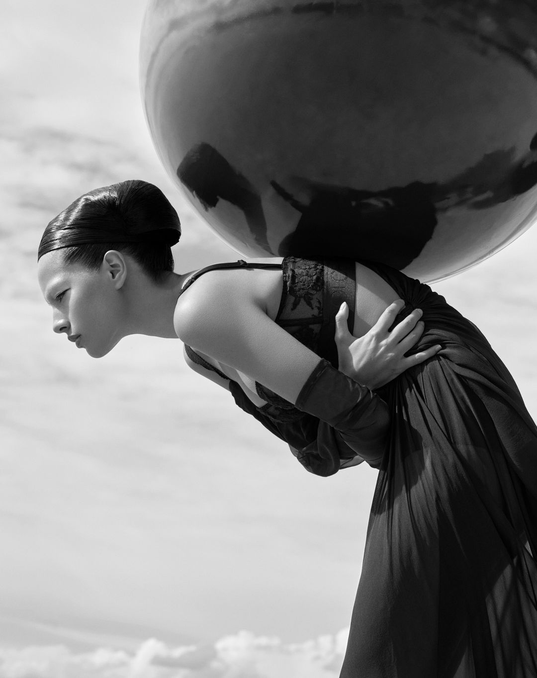 Dramatic monochrome fashion shot of an adult woman in a dress bent over under the weight of a black sphere against a cloudy sky.