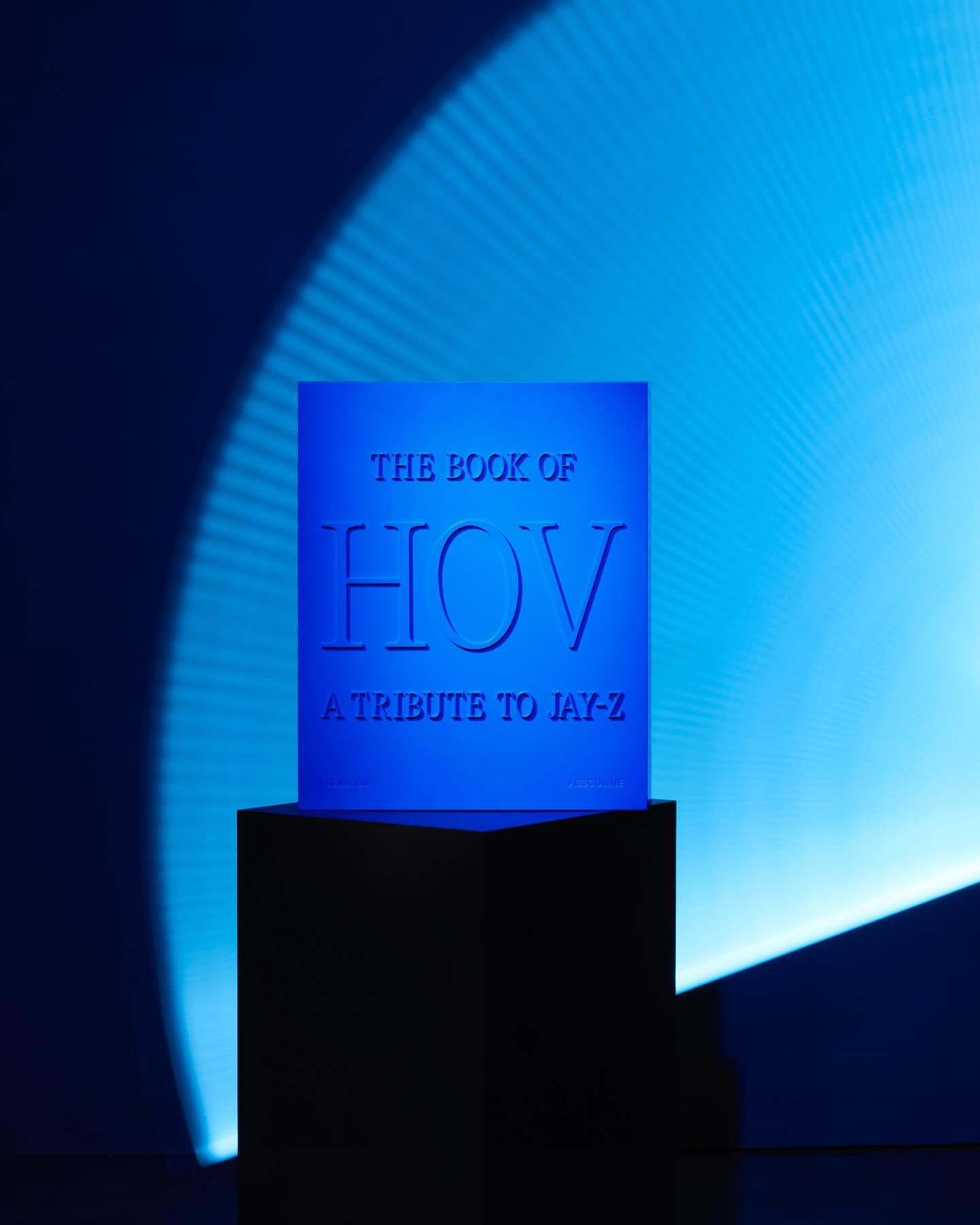 Close-up of a blue promotional cube on a black base with the embossed title "The Book of HOV, A Tribute to Jay-Z," illuminated by blue lighting.