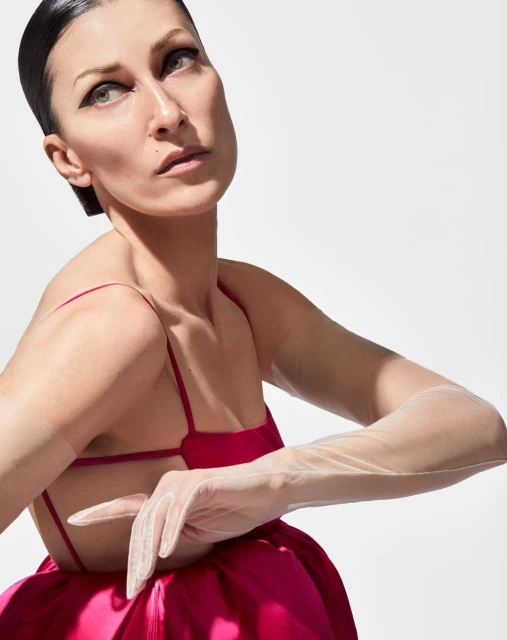 Woman in a pink dress and sheer gloves poses against a white backdrop, her gaze directed upwards, in a studio fashion portrait.