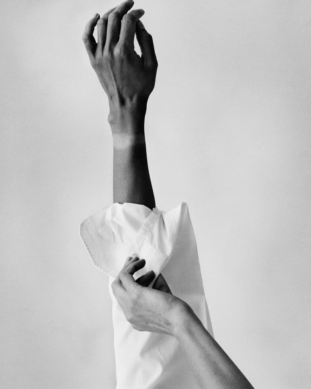 Black and white photo of fair-skinned adult female with raised arm, wearing a white shirt and tan line, in a neutral indoor setting.
