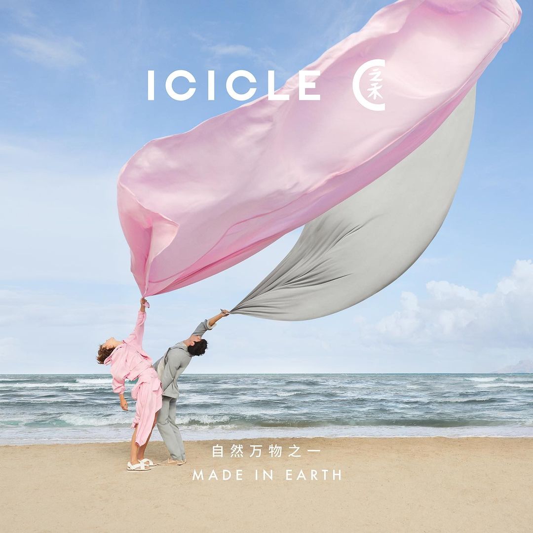 Two people in coordinated outfits hold a flowing pink and gray fabric on a sandy beach against a backdrop of ocean and blue sky.