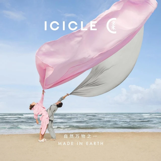 Two people in coordinated outfits hold a flowing pink and gray fabric on a sandy beach against a backdrop of ocean and blue sky.