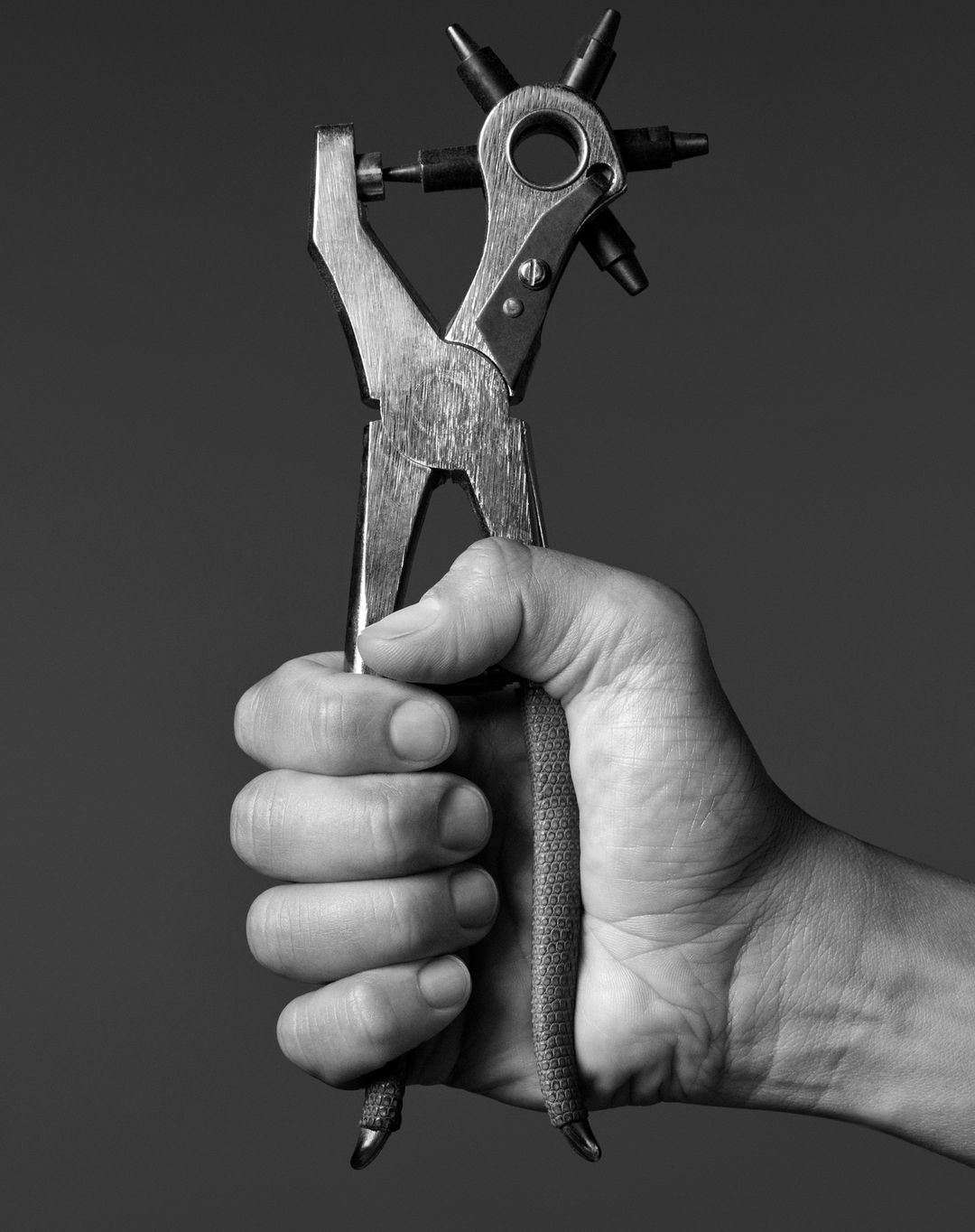 Close-up of a hand holding a rotary leather punch against a dark background. The tool is silver with a black punch selector.