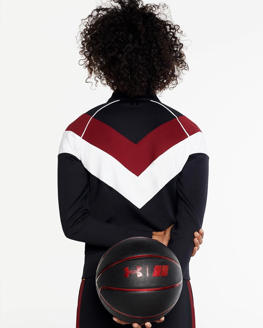 Rear view of a woman with curly hair, wearing a track jacket, holding a black basketball against a white backdrop in a studio shot.