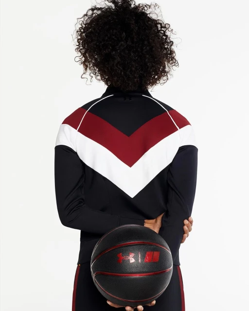 Rear view of a woman with curly hair, wearing a track jacket, holding a black basketball against a white backdrop in a studio shot.
