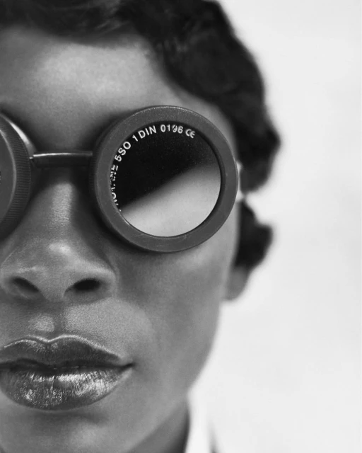 Close-up black and white portrait of an adult woman with sunglasses and dark, wavy hair, looking forward against a white background.