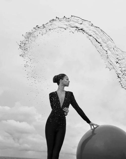 Elegant woman in black dress poses near large red sphere under a splash of water against a cloudy sky in a black and white outdoor shot.