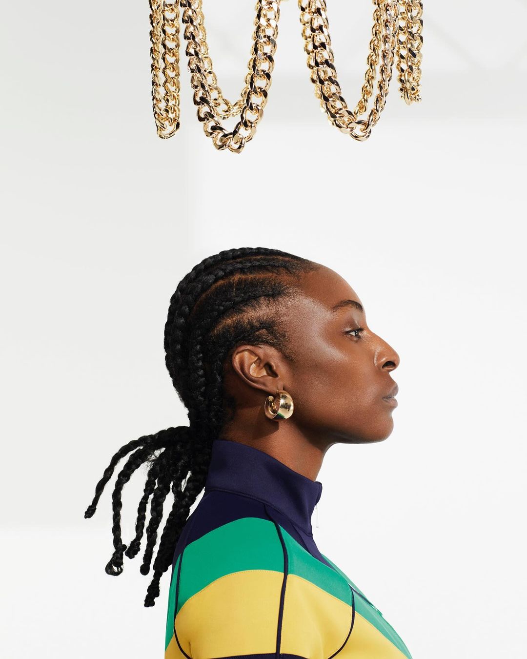 Profile shot of a Black woman with braided hair wearing a color-blocked top and gold hoops, with gold chains hanging above against white.