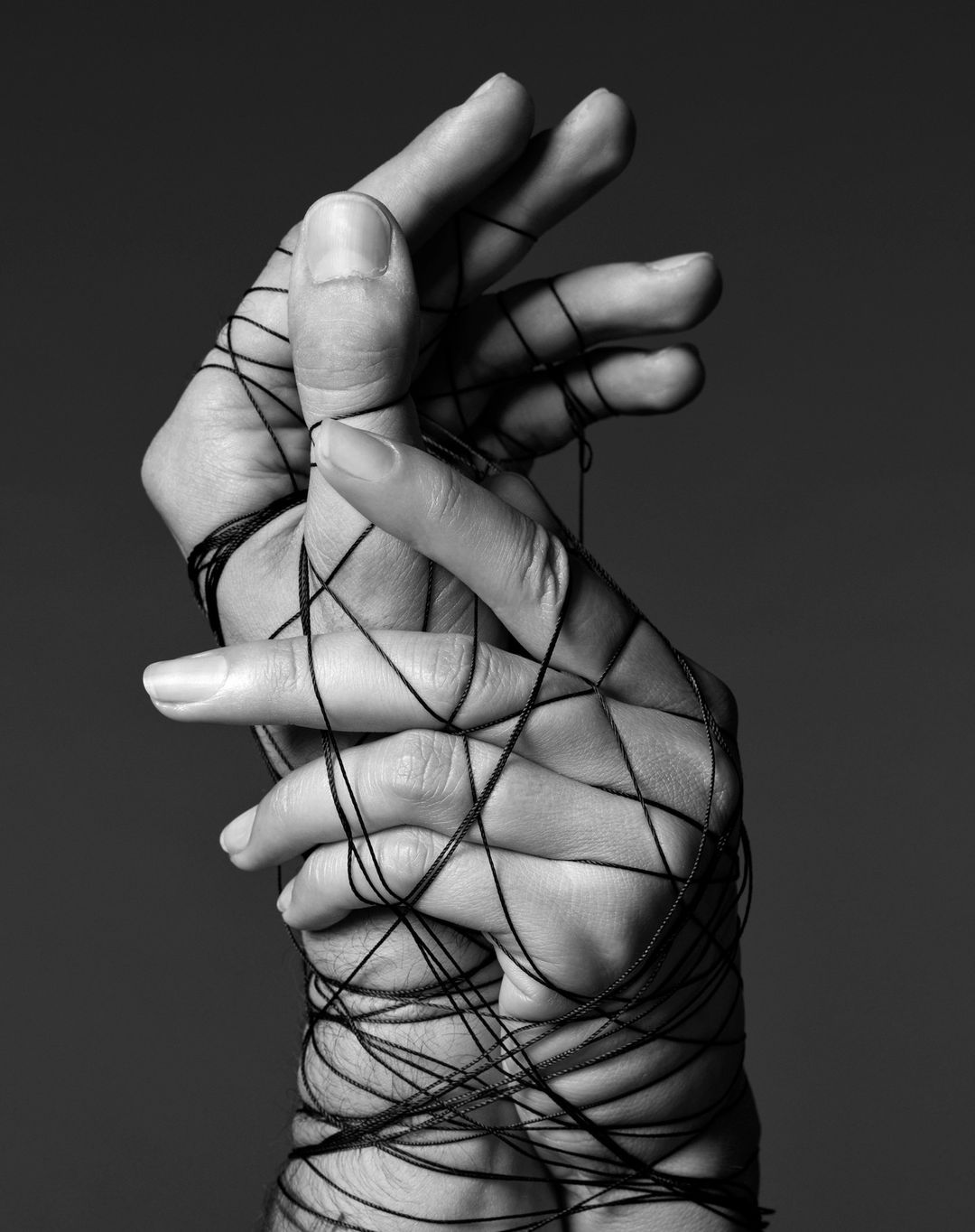 Two hands intertwined with black string against a dark grey background in a studio setting. The black and white photo has an editorial style.