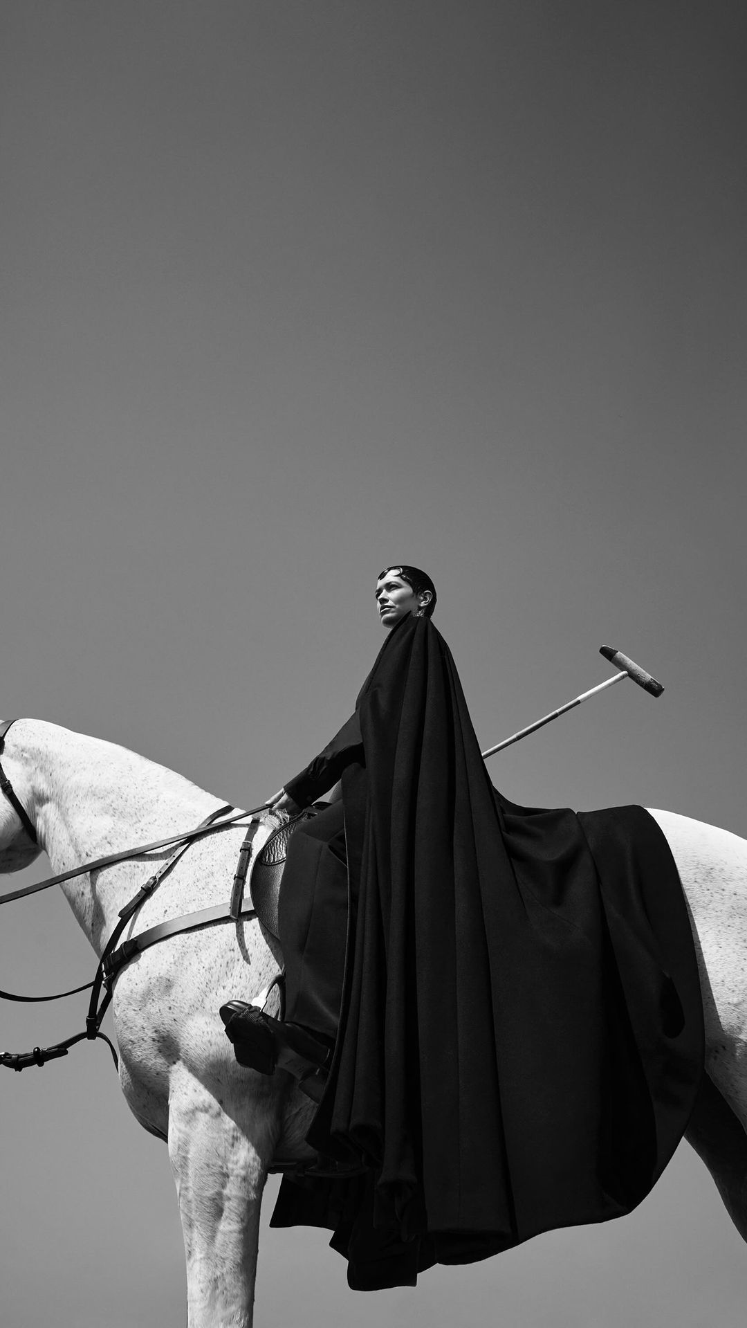 Black and white portrait of a woman in a cape riding a white horse outdoors in natural light against a clear, light sky.