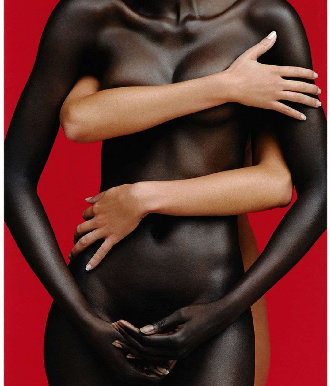 Two nude women embrace in a studio shot with a red background. One woman is painted black. Sensual studio shot with fine art aesthetic.