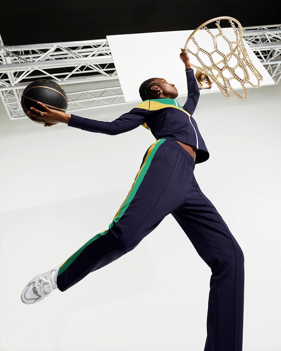 Athletic woman in navy track suit jumps to make a basket with a basketball in studio against a white and gray background.