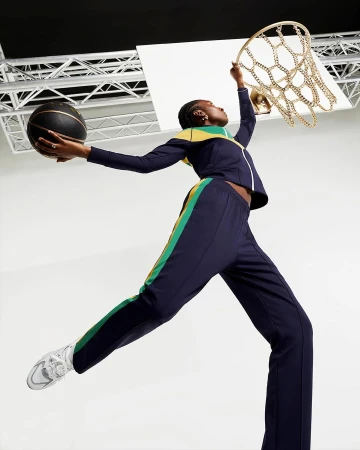 Athletic woman in navy track suit jumps to make a basket with a basketball in studio against a white and gray background.