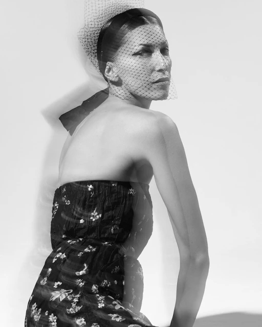Black and white portrait of a woman in a floral dress and a veiled headpiece looking over her shoulder in a photo studio setting.