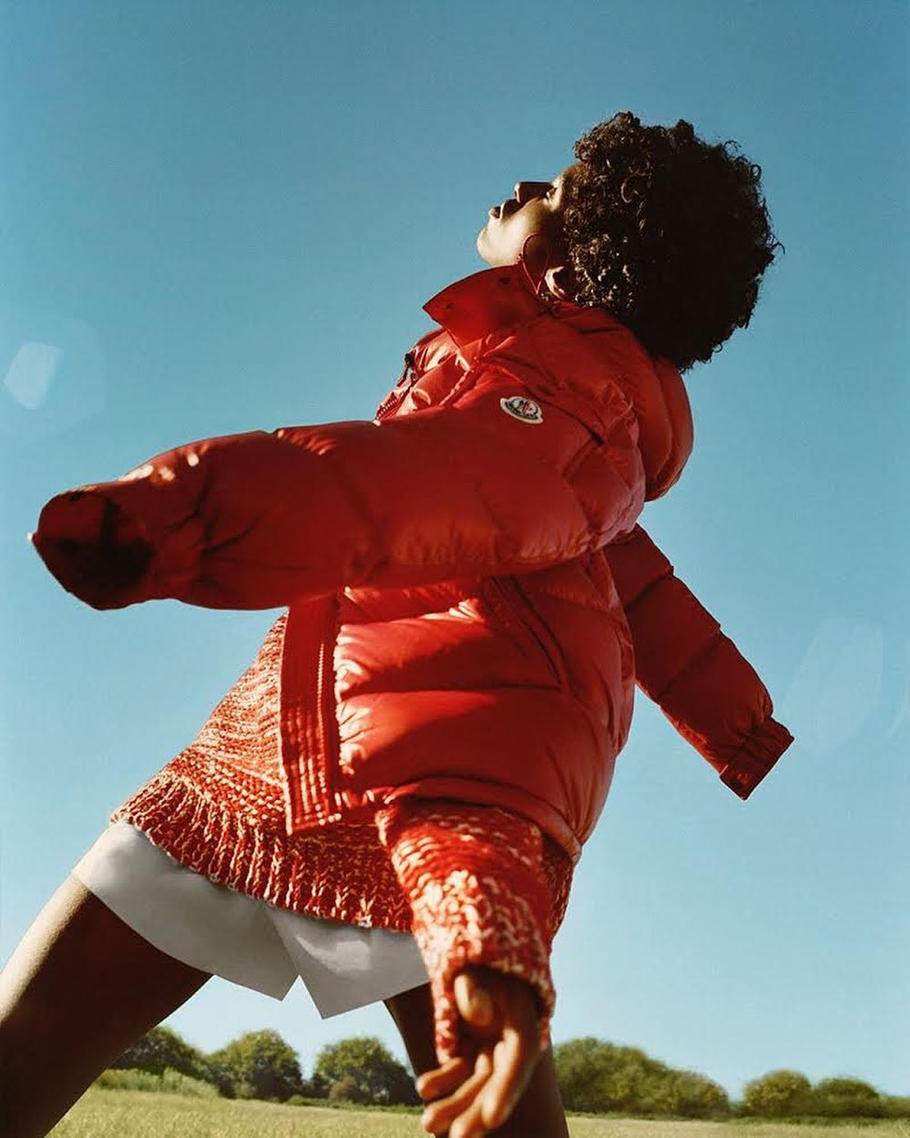 Woman in red puffer jacket, white shorts, and curly hair standing in a field under a clear blue sky, looking up in an outdoor setting.