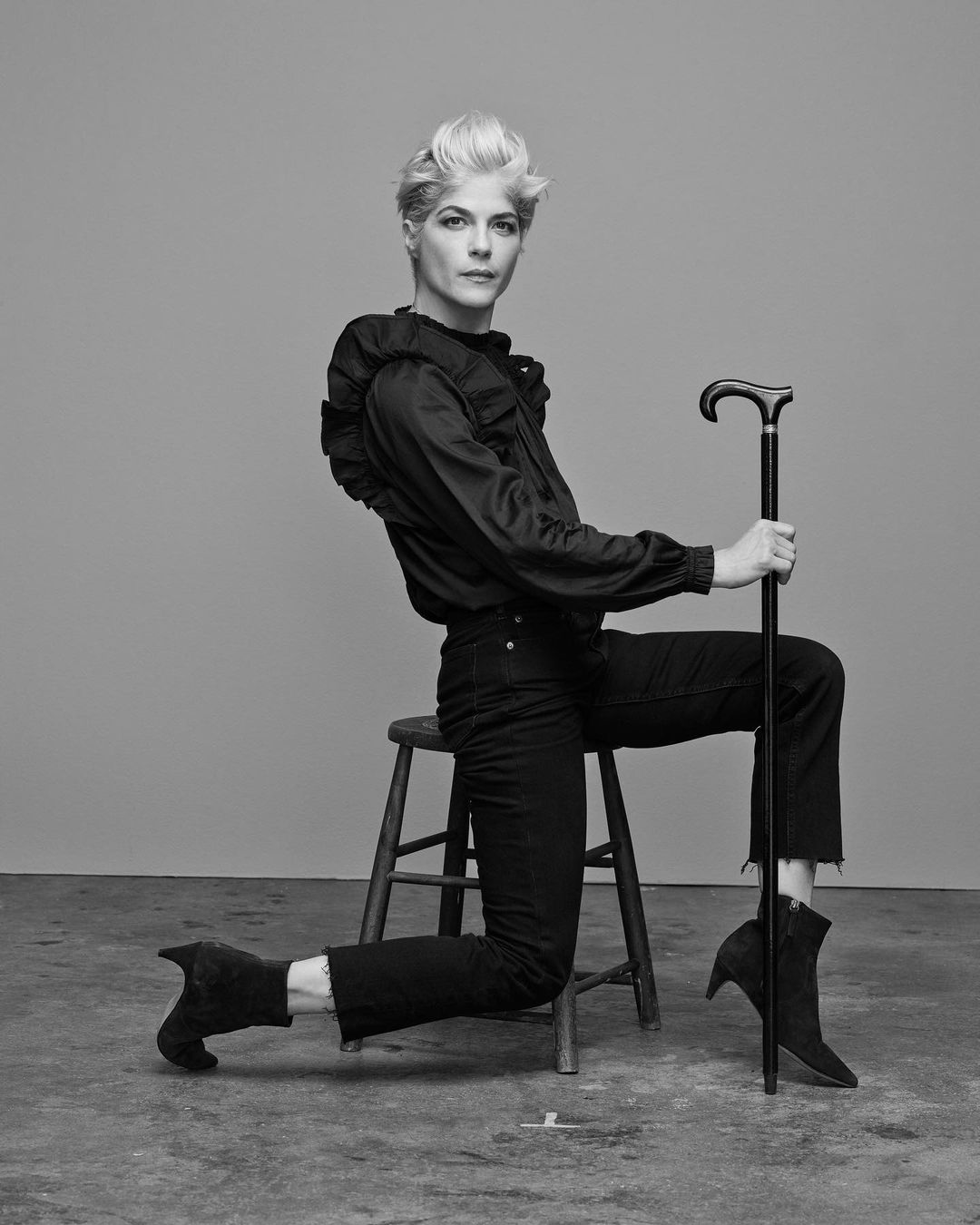 Black-and-white portrait of a blonde woman kneeling on one knee on the floor while seated on a stool and holding a cane.