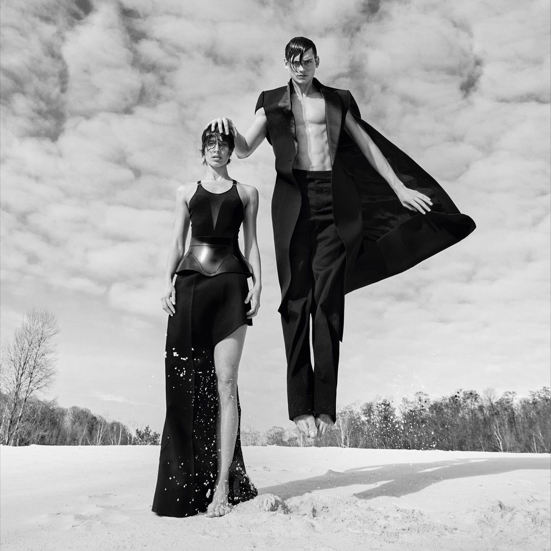 Black and white portrait of a woman standing and a man levitating in black attire against a cloudy sky