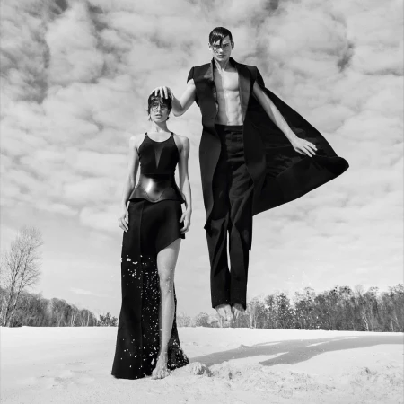 Black and white portrait of a woman standing and a man levitating in black attire against a cloudy sky