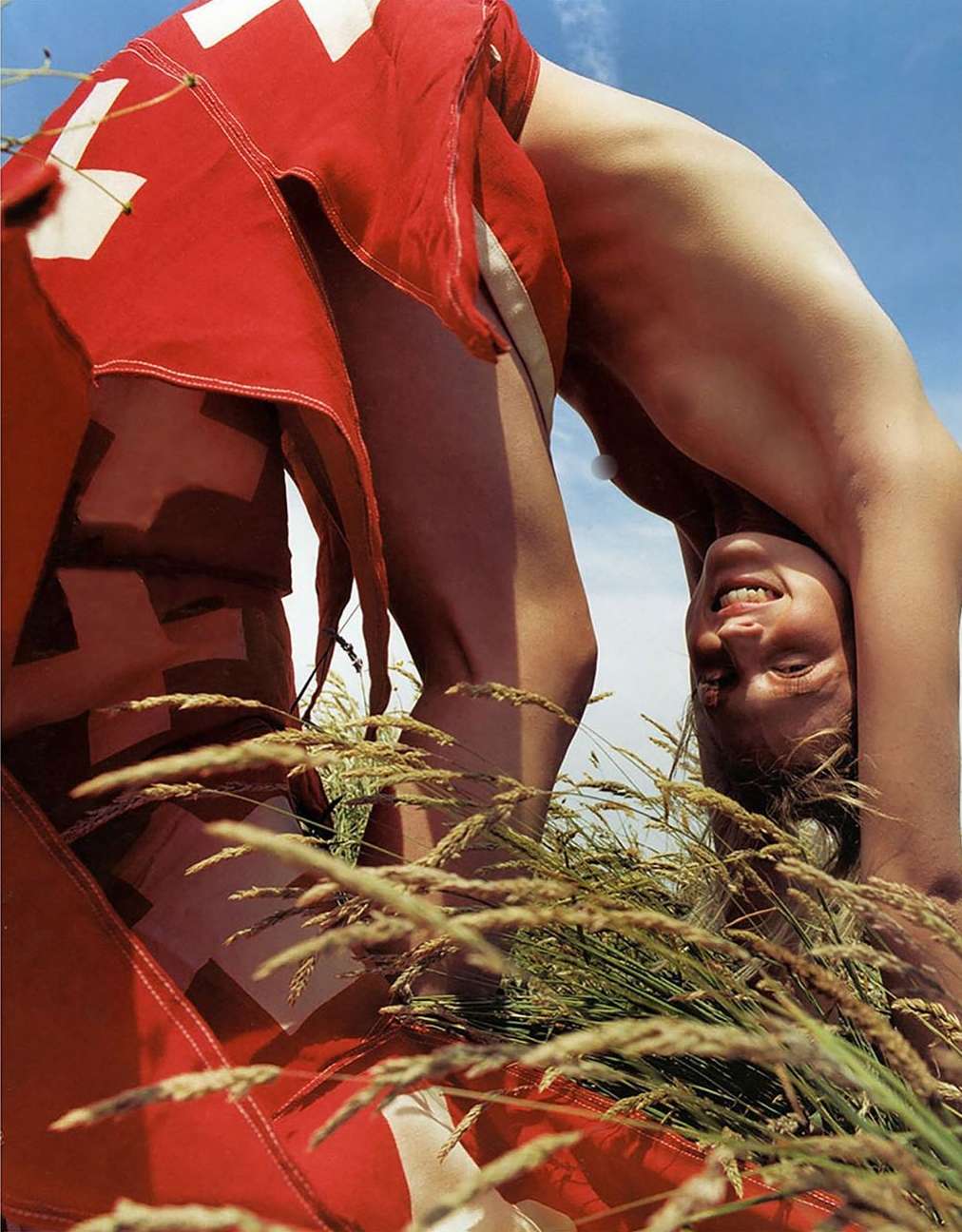 Adult female in arched pose wearing a red and white flag, outdoors in grass, set against blue sky.