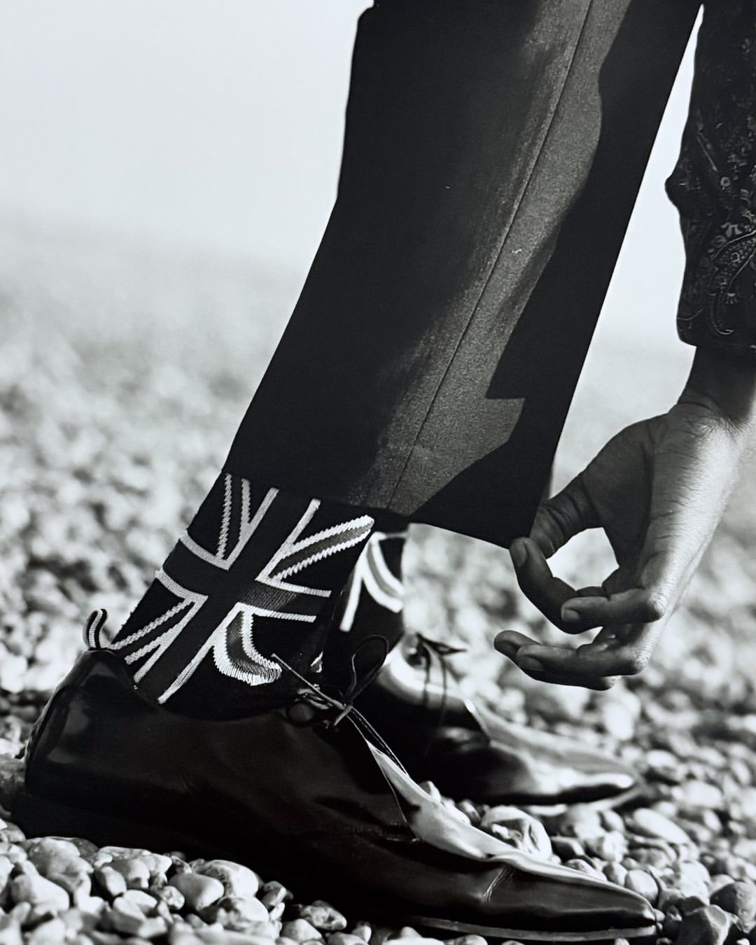 Low angle, black and white close-up of person adjusting trouser leg, revealing socks with a Union Jack pattern and black leather shoes.