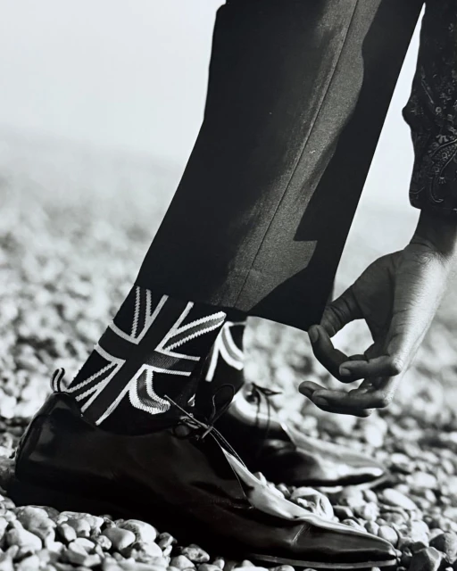 Low angle, black and white close-up of person adjusting trouser leg, revealing socks with a Union Jack pattern and black leather shoes.
