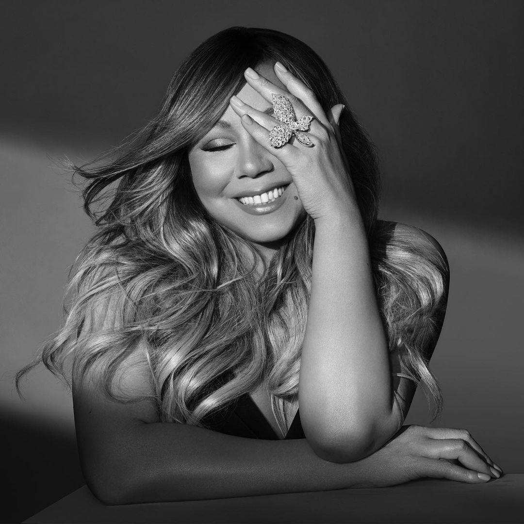 Monochrome portrait of Mariah Carey, eyes closed, diamond butterfly ring, long wavy hair, hand on face, resting arm.