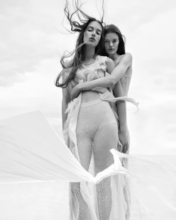 Monochrome fashion portrait of two female models in textured sheer outfits with long hair blowing in wind against an open sky.