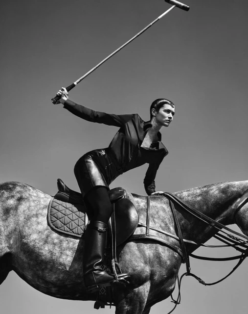 Black and white shot of woman on horseback holding polo mallet outdoors, set against a clear sky, capturing sport and elegance.