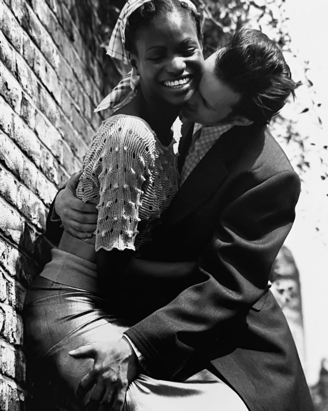 A smiling African-American woman in a light-colored sweater is being embraced and kissed by a light-skinned man outdoors near a brick wall.