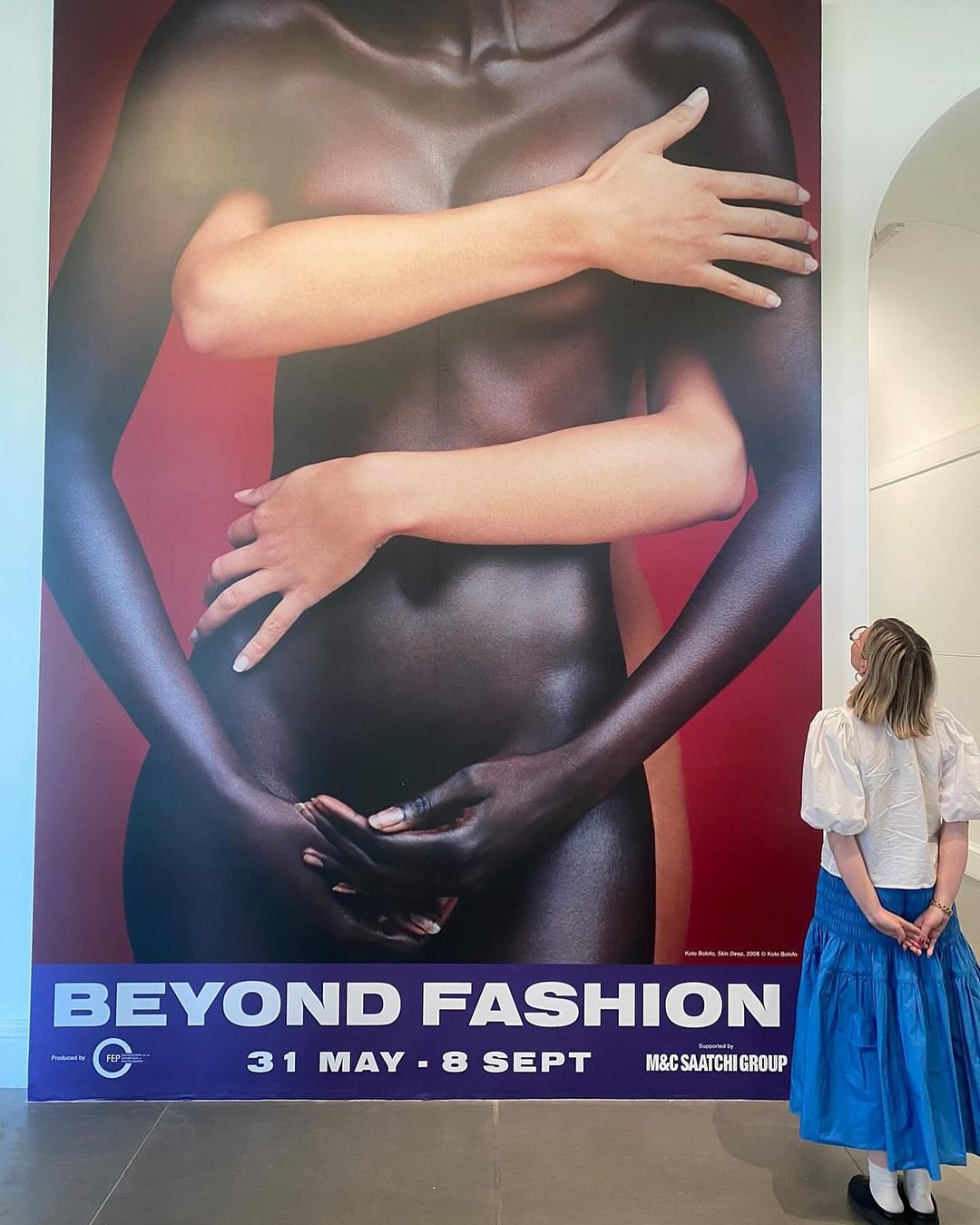 Woman views the Beyond Fashion art exhibition poster featuring two women's torsos with diverse skin tones and interlocked hands in a gallery.