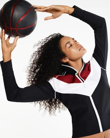 Woman with curly dark hair wearing a color-blocked zip jacket holding a black and red basketball above her head.