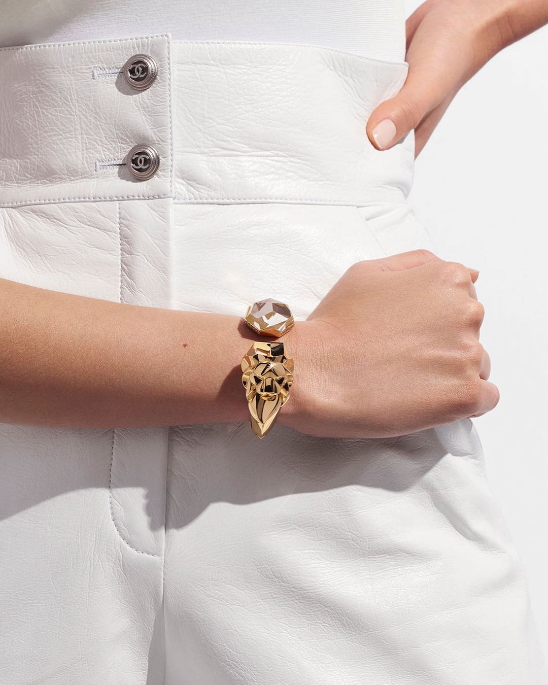 Closeup of a woman's wrist featuring a large, ornate gold cuff bracelet, set against a backdrop of white leather apparel.