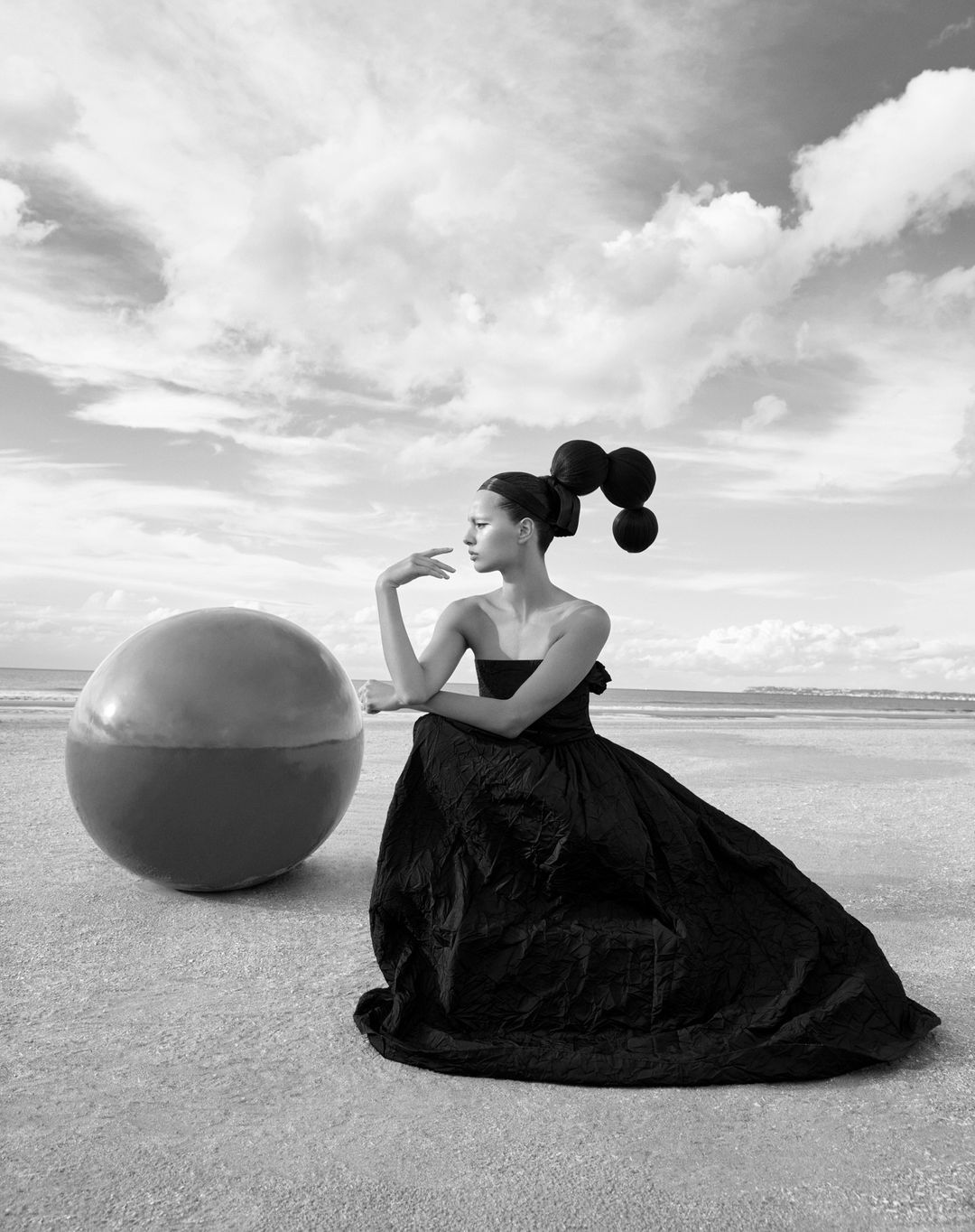 Monochrome portrait of a woman in a black gown, artistically posed with a sphere against a sky and beach backdrop in black and white.