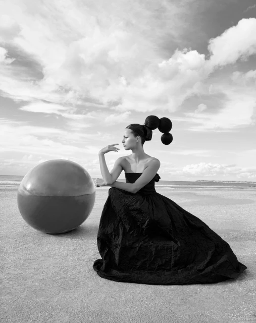 Monochrome portrait of a woman in a black gown, artistically posed with a sphere against a sky and beach backdrop in black and white.