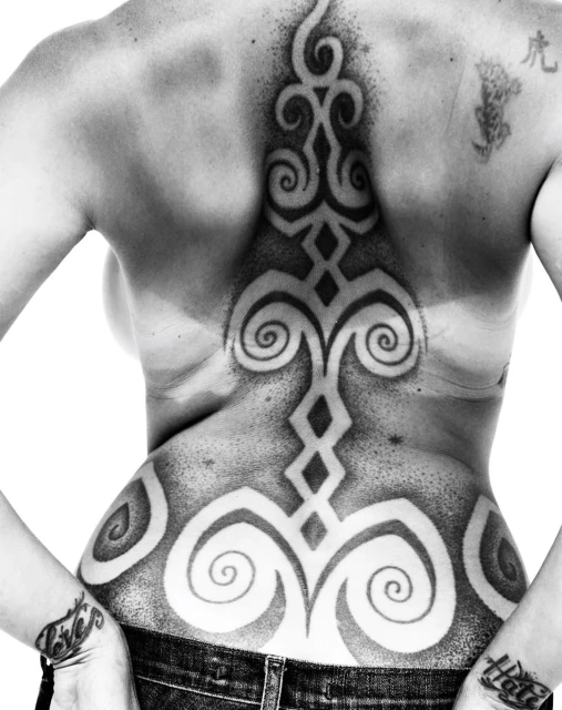 Black and white shot of an adult female's back featuring tribal-style tattoo designs, seen from the rear view with hands on hips.